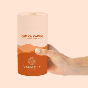 A hand holds a tall, orange and tan container of VANADEY AAP KA KADHA Premium Organic Herbal Infusion.